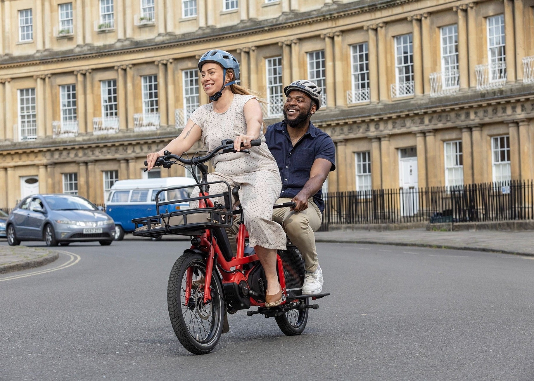 Two people riding a red cargo electric bike on a city street with classical architecture in the background.