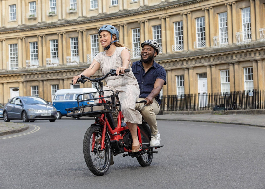 Two people riding a red cargo electric bike on a city street with classical architecture in the background.