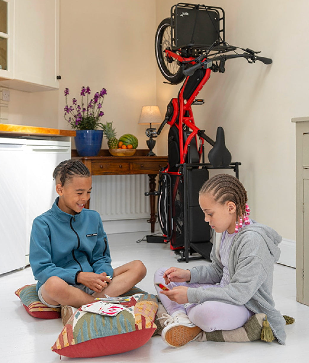 Two children sitting on a kitchen floor with a bicycle mounted on a stand in the background.