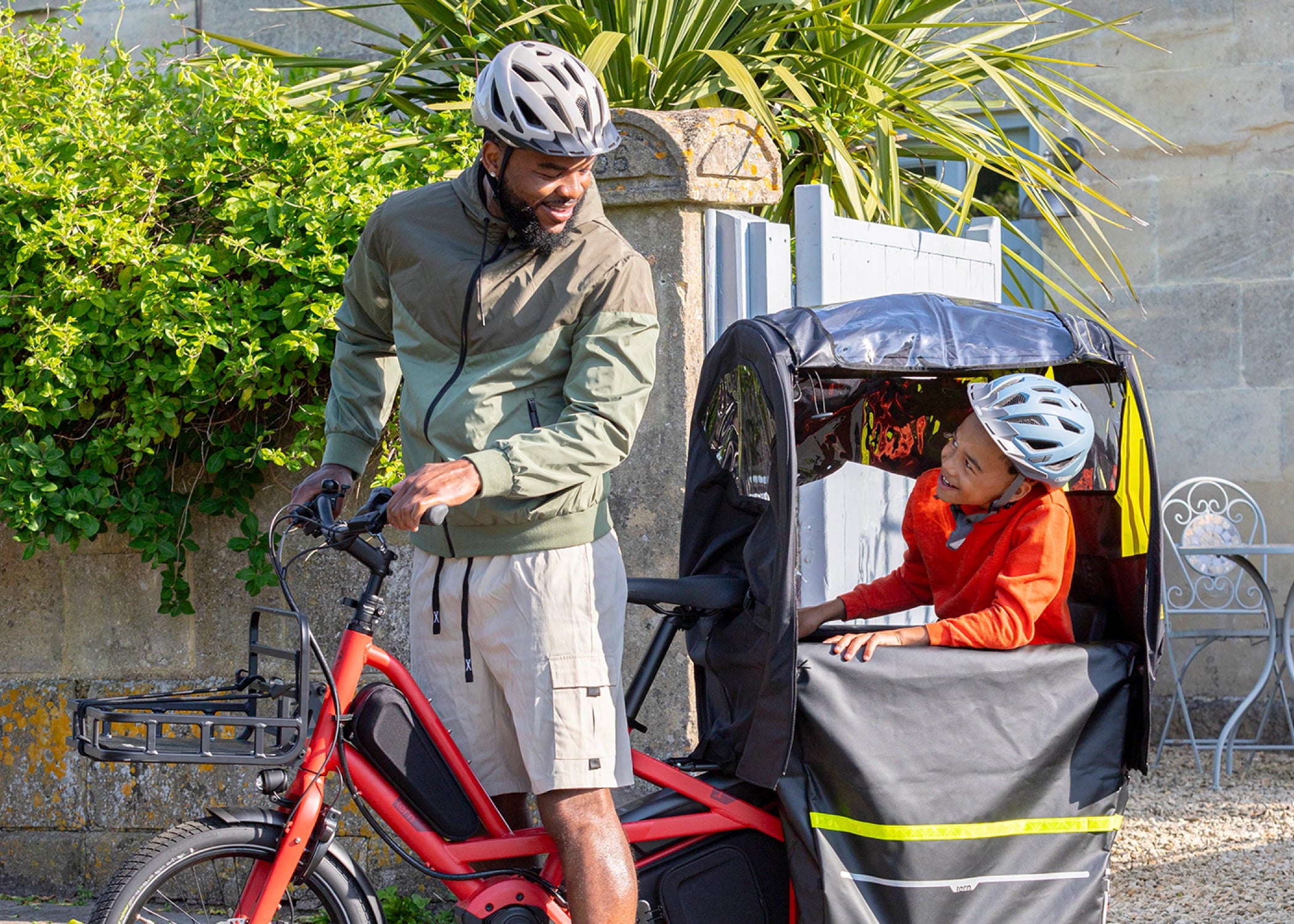 Man with child in a bike trailer outdoors near greenery and a stone wall.