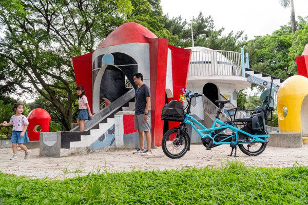 A bright turquoise electric cargo bike with a rear cargo box is parked in front of a colorful children's playground with red, white, and yellow structures. Two children and an adult are standing near the playground, and trees and grass are visible in the background.