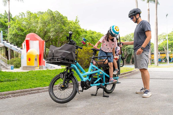 Blue Cargo bicycle with a child on it and a person standing next to it in the park setting.