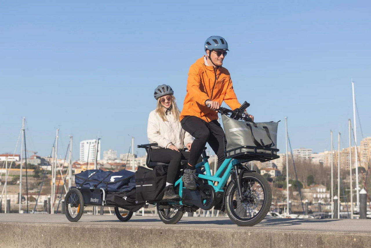 Two people riding a Tern cargo bike with a trailer on a waterfront path.