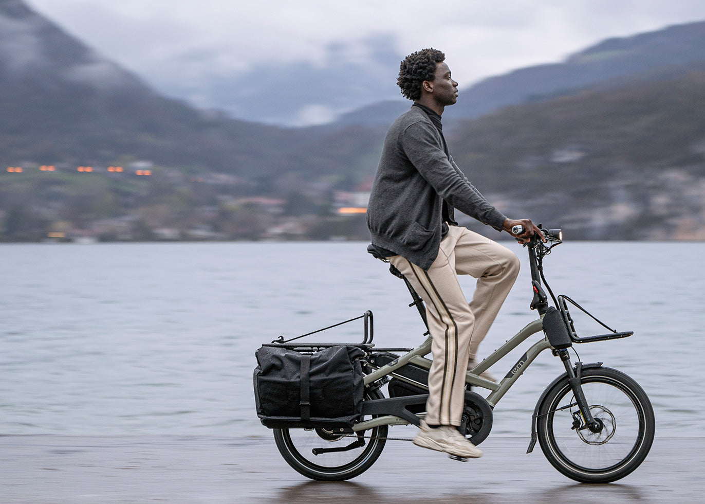 a man riding a cargo bicycle with a basket on a lake with mountains in the background