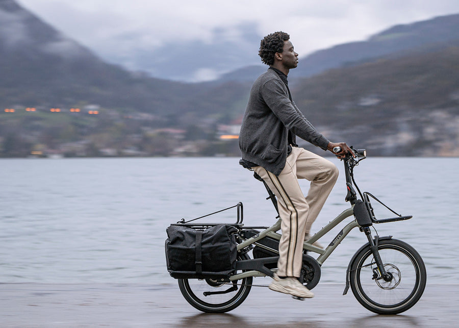 a man riding a cargo bicycle with a basket on a lake with mountains in the background