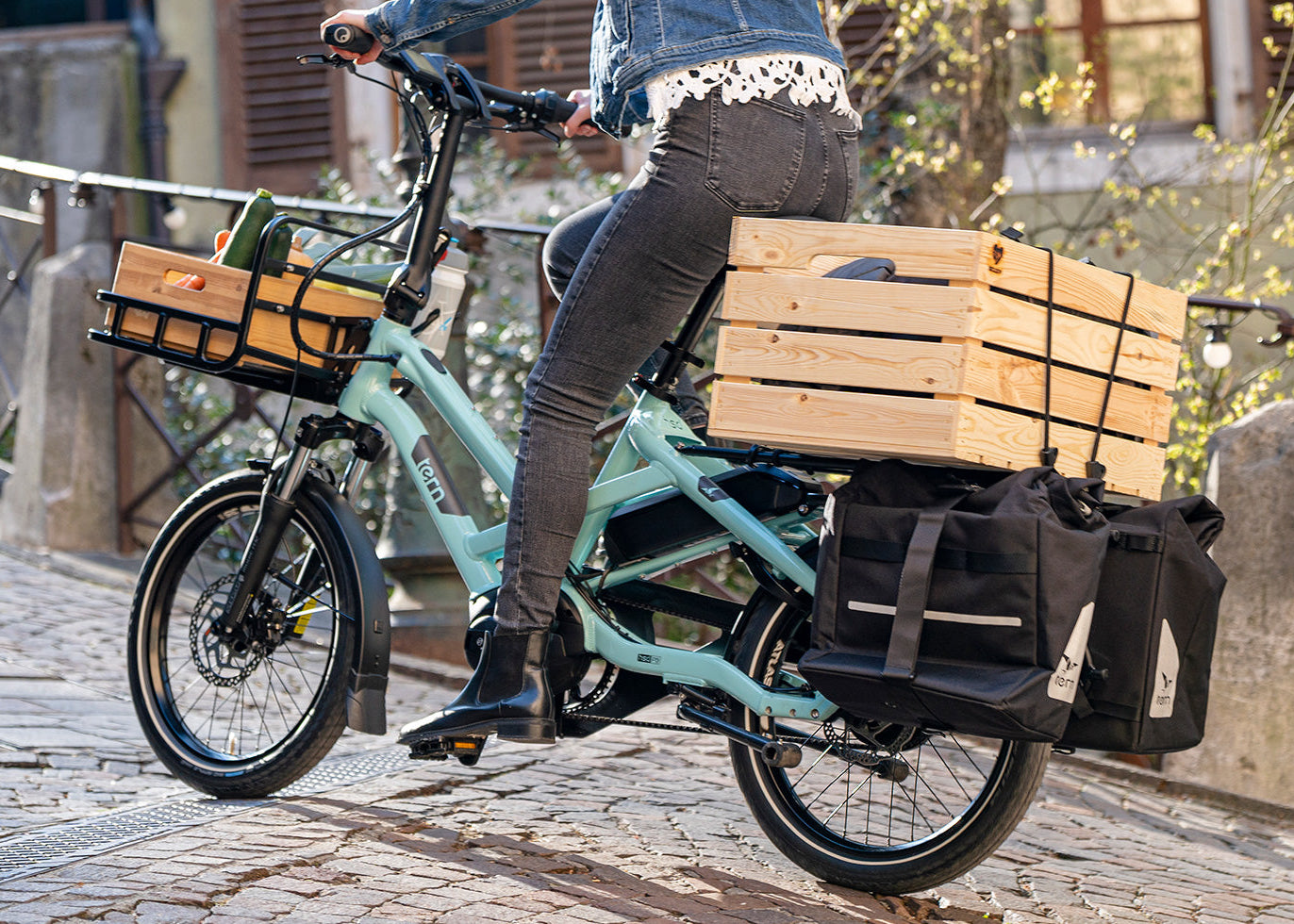 Person riding a teal cargo bike with wooden baskets on a cobblestone street.