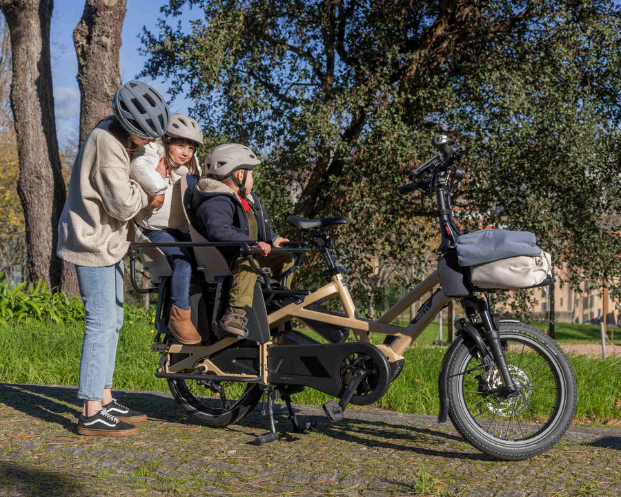 A family of four enjoys a sunny day on a beige cargo electric bicycle with a rear passenger seat, parked in a park. Two adults wear helmets and jackets while standing next to the bike, and two children sit on the rear rack wearing helmets and jackets.