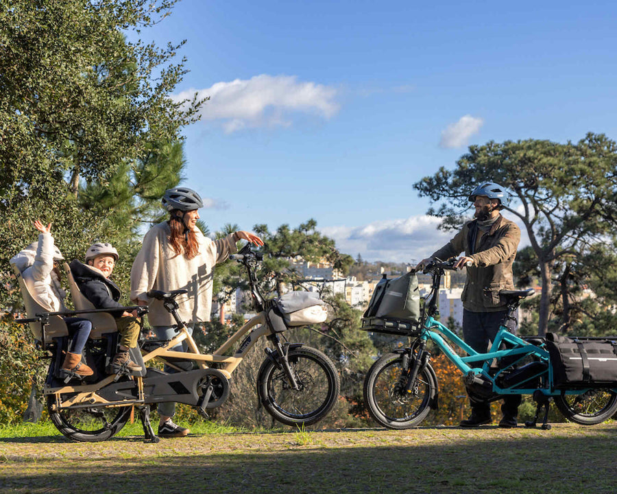 A family of four with two electric Tern bicycles in an outdoor park setting under a blue sky. One bicycle has a large rear cargo bed with a child sitting in it, while the other is a turquoise utility bike with a front basket.